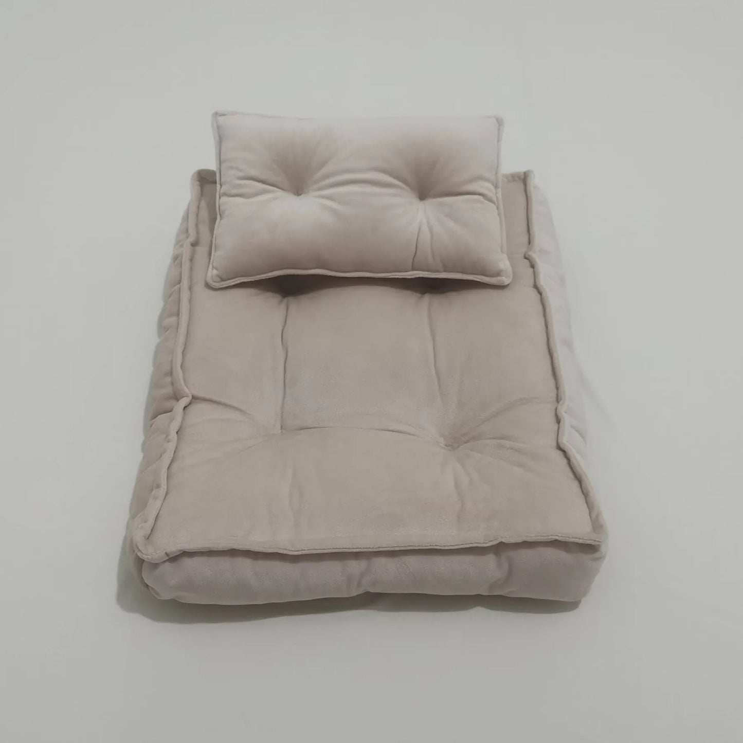 A plush beige pet bed with a matching cushioned pillow, both tufted for added comfort, placed on a smooth white surface.