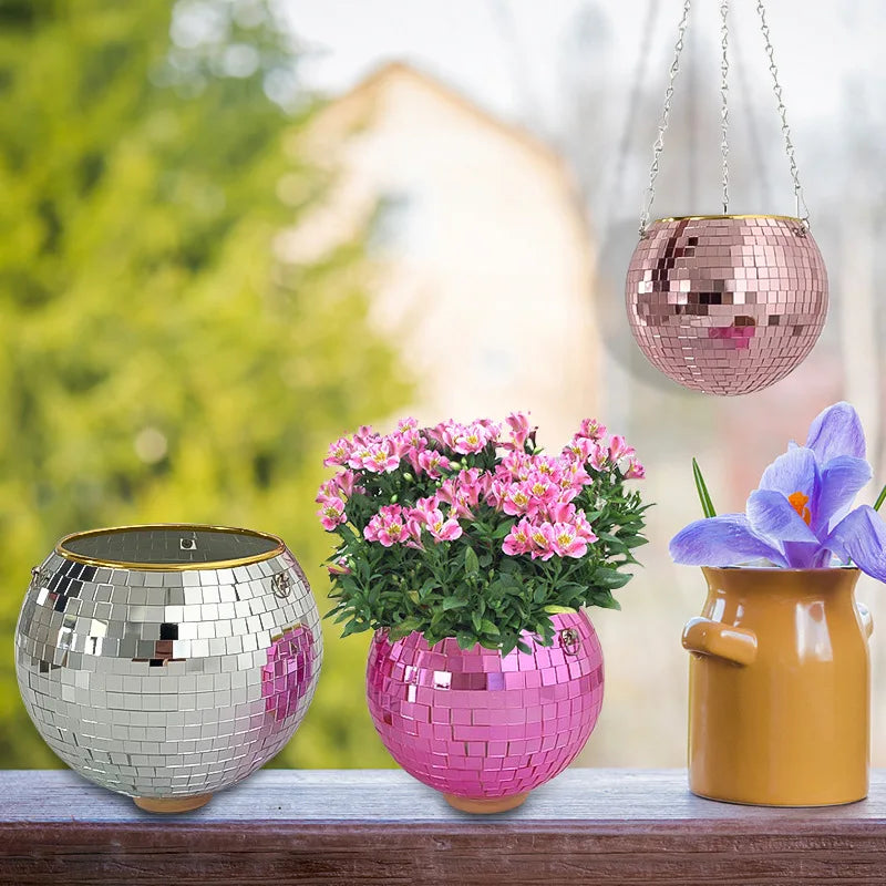 Three disco ball planters, one silver, one pink with flowers, and a pink hanging planter, alongside a purple flower in a yellow jug on a wooden surface.