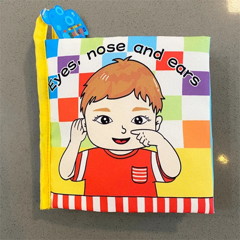 Children's cloth book cover with a cartoon child in a red shirt touching nose and ears. The title reads "Eyes, nose and ears" on a colorful checkered background.