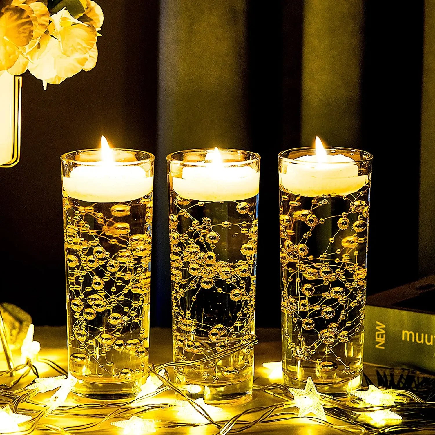 Three tall glass candles with floating wax discs and intricate bubbles are lit, surrounded by decorative string lights and part of a book, creating a warm, cozy ambiance.