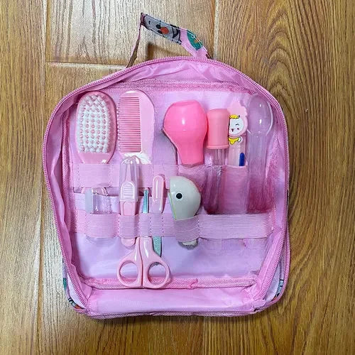 Pink baby grooming kit with clear compartments, containing a hairbrush, comb, medicine dropper, nasal aspirator, scissors, nail clipper, and file, on a wooden floor.