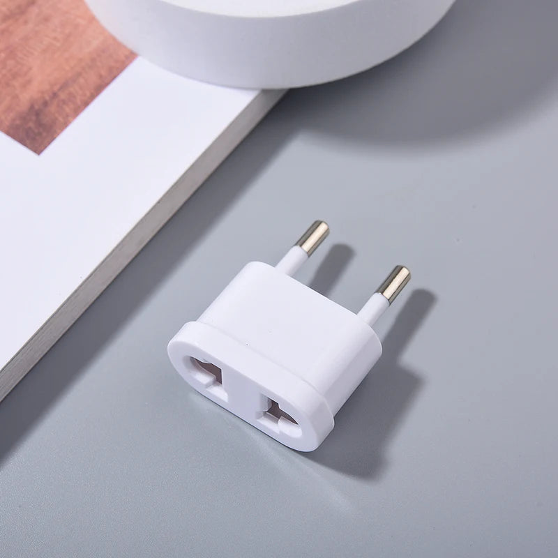 A white plug adapter with two round metal prongs lies on a light gray surface. A book and a circular object are partially visible in the background.