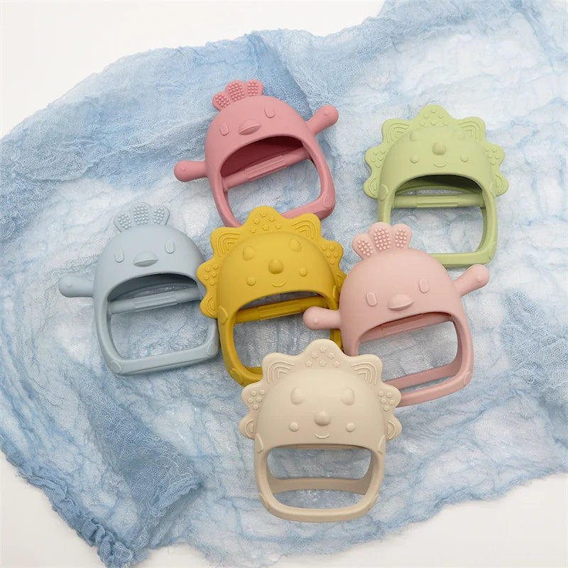 Colorful silicone baby teethers shaped like cartoon characters are arranged on a soft blue cloth. The teethers are in pastel shades of blue, pink, green, yellow, and beige.