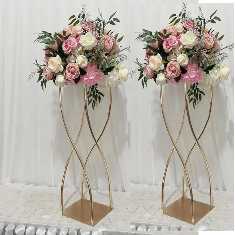 Elegant floral arrangements with pink and white roses, peonies, and greenery atop gold spiral stands against a white curtain backdrop on a white petal-covered surface.