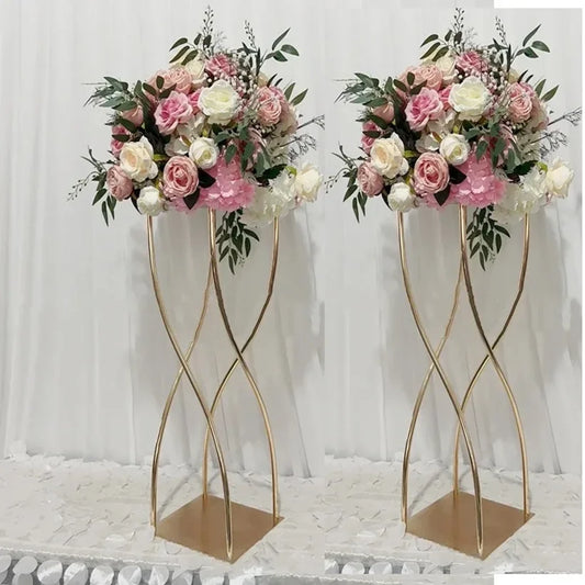 Elegant floral arrangements with pink and white roses, peonies, and greenery atop gold spiral stands against a white curtain backdrop on a white petal-covered surface.
