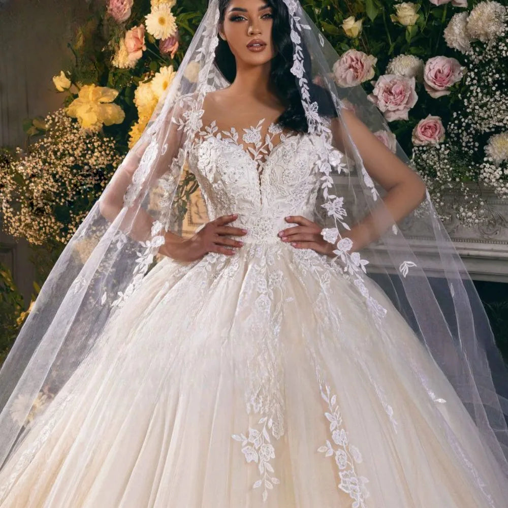 Bride in a lace bridal gown with floral appliqué and a long veil poses in front of a lush floral backdrop featuring white and pink roses with greenery.