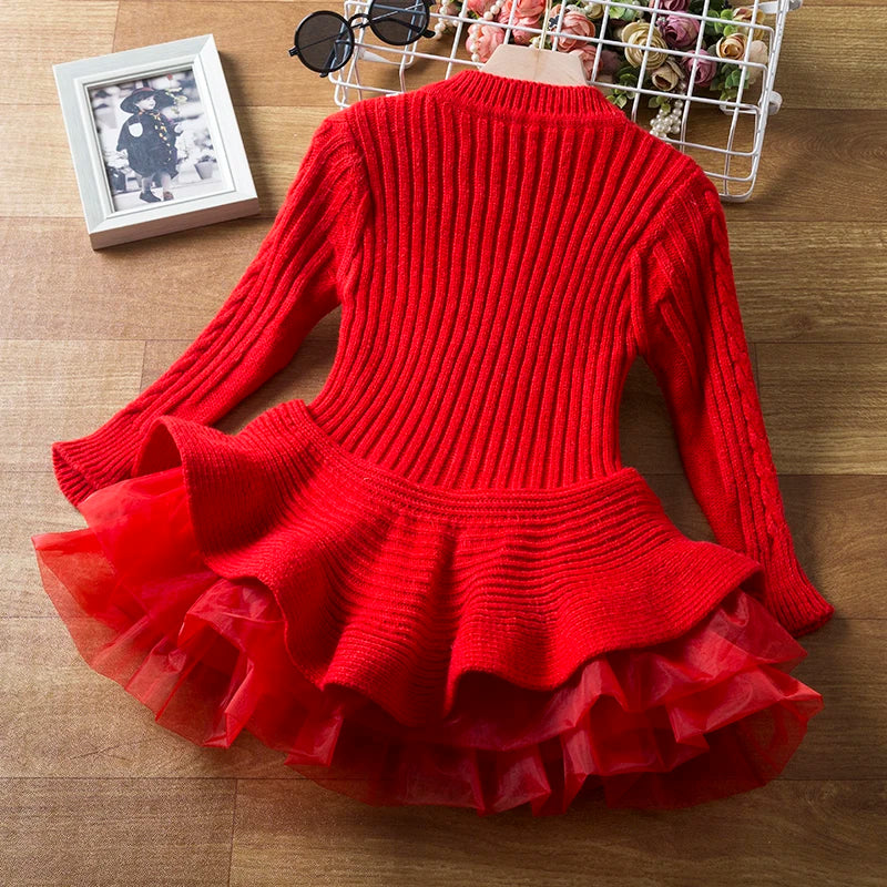 A vibrant red sweater dress with a ribbed knit top and ruffled tulle skirt lies on a wooden floor. Adjacent are a pair of black sunglasses, a flower basket, and a monochrome photo.