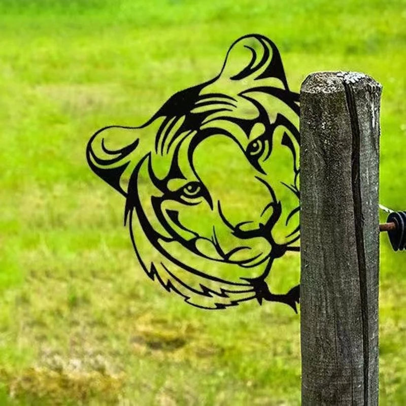 A metal artwork of a tiger's face is attached to a wooden post in a grassy field. The intricate design features bold lines creating the tiger's eyes, nose, and stripes.