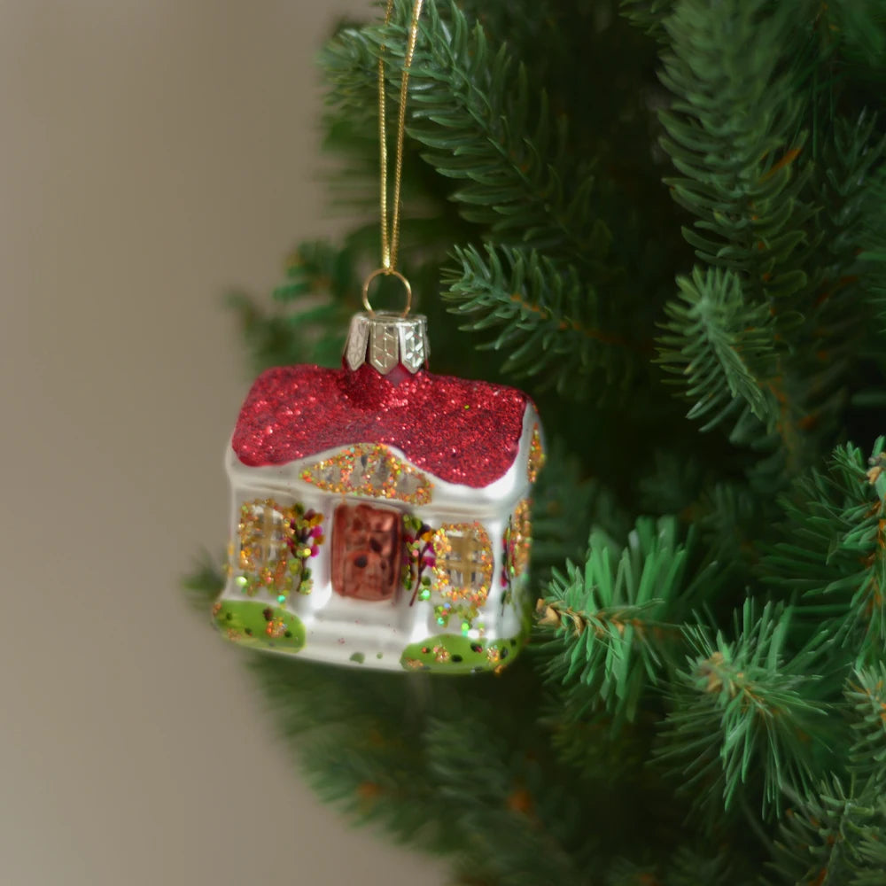 Christmas tree ornament shaped like a house with a glittery red roof, gold details, and adorned windows, hanging from a green pine branch.
