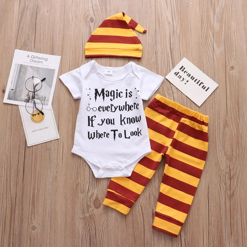 Striped yellow and red baby outfit includes a hat, pants, and a onesie with the text "Magic is everywhere if you know where to look," on a wooden surface with booklets nearby.