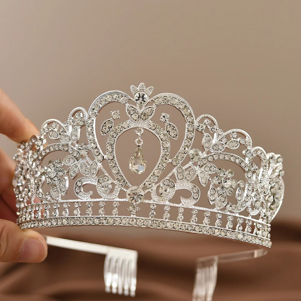 A silver tiara adorned with intricate floral patterns and sparkling gemstones, featuring a central teardrop crystal. A hand holds the tiara against a neutral background.