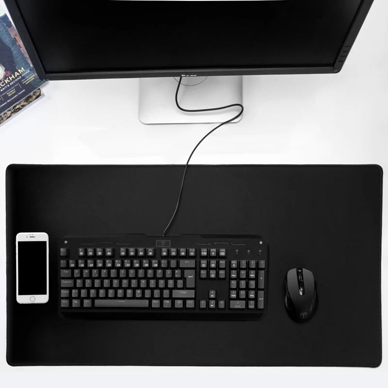 A black keyboard and mouse on a dark desk mat, with a smartphone on the left. A monitor is in the background with a visible power cord, next to a book on the desk.