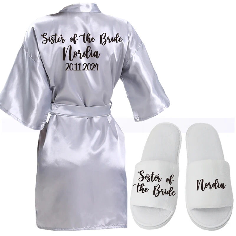 Silver satin robe with "Sister of the Bride" and "Nordia 20.11.2024" printed on the back, paired with white slippers labeled "Sister of the Bride" and "Nordia."