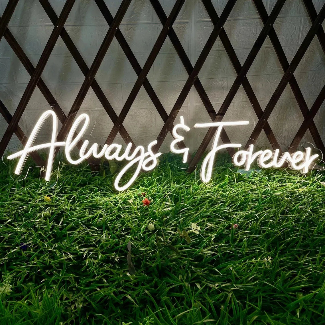 Neon sign reading "Always & Forever" in cursive, placed on lush green grass against a wooden lattice backdrop, creating a vibrant and romantic atmosphere.
