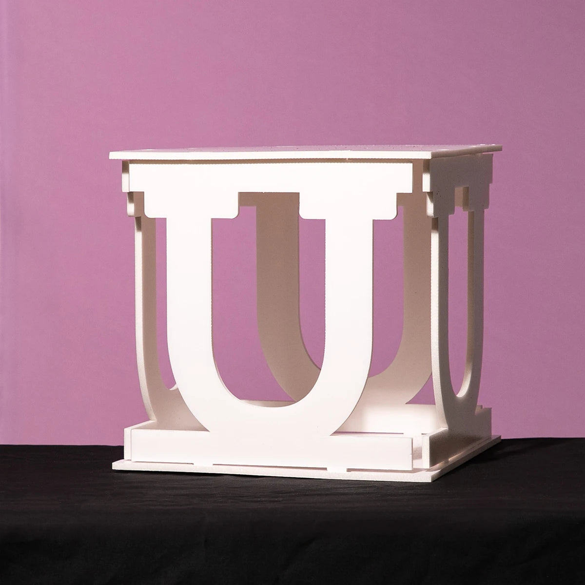 White cubic structure with U-shaped cutouts on all sides, set against a purple background and resting on a black surface.