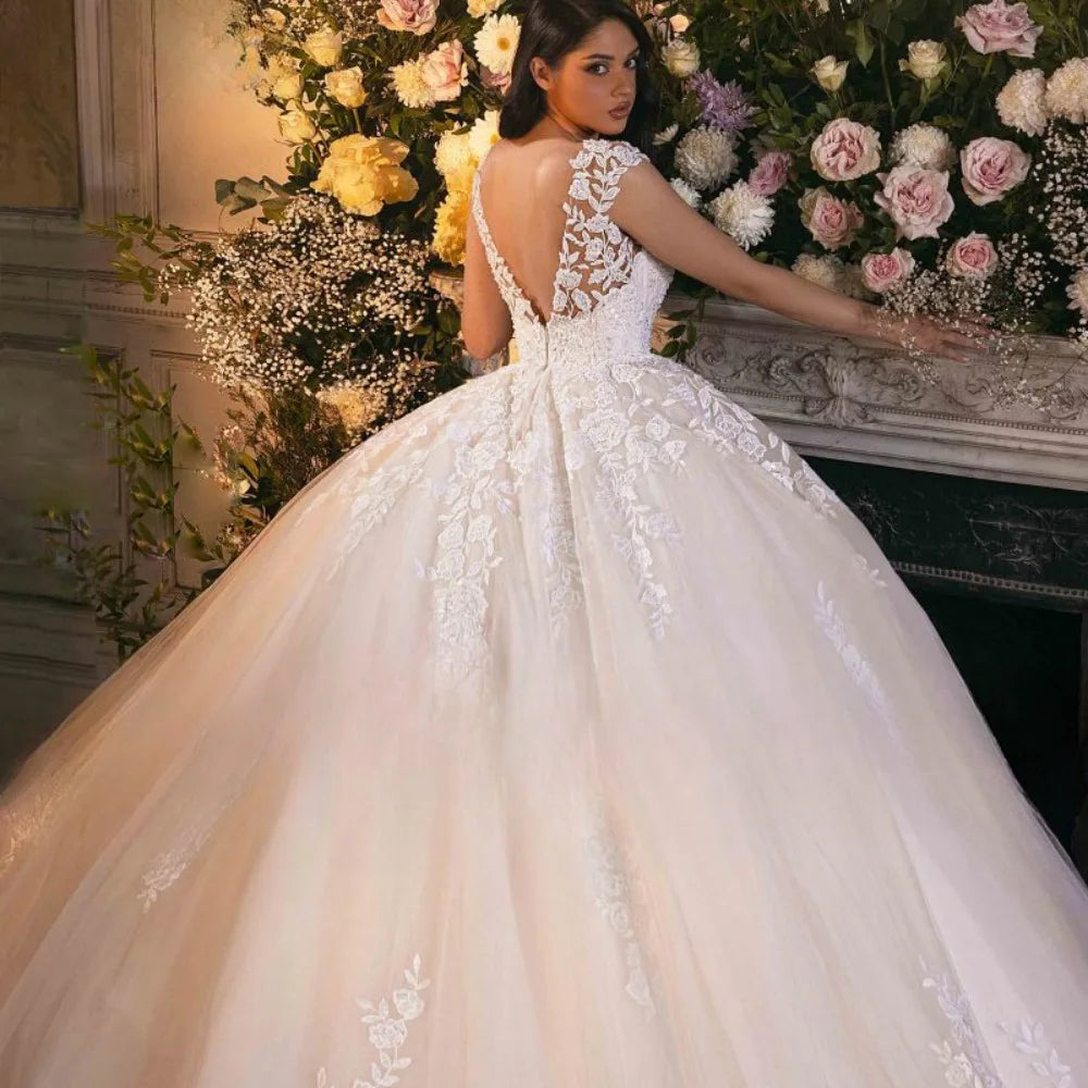 Bride in a voluminous white gown with floral lace details stands beside a floral arrangement. The dress features a low back and intricate lace embellishments.