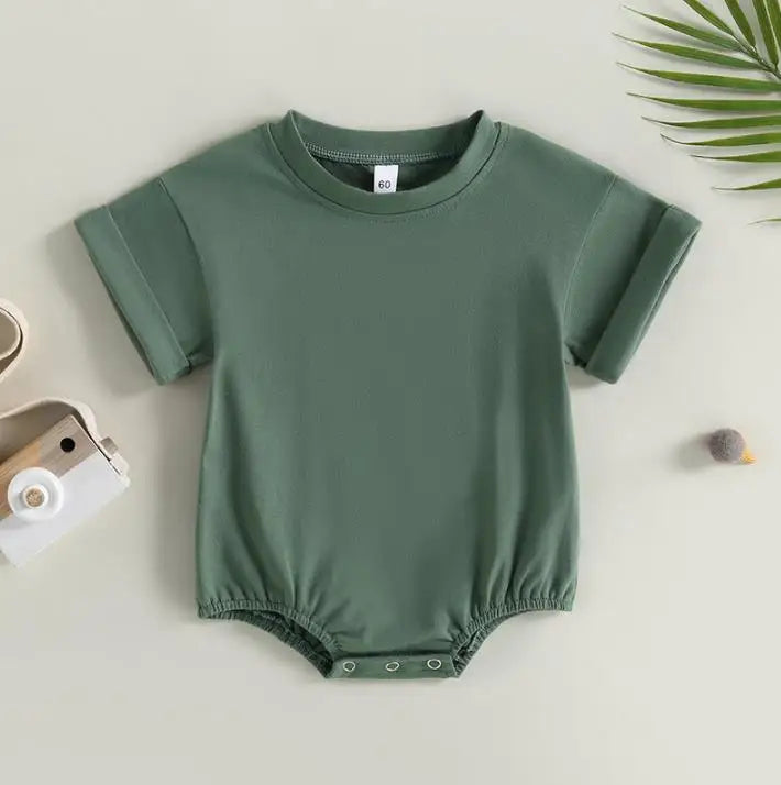 Green baby romper with short sleeves and snap buttons at the bottom, displayed flat. Nearby, a toy car, a small ball, and a green leaf for decoration.