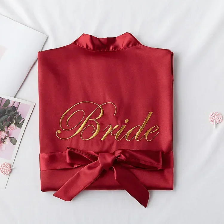 A red satin robe with "Bride" embroidered in gold on the back. It is neatly folded with a tied sash, set against a white background with decorative pink elements.
