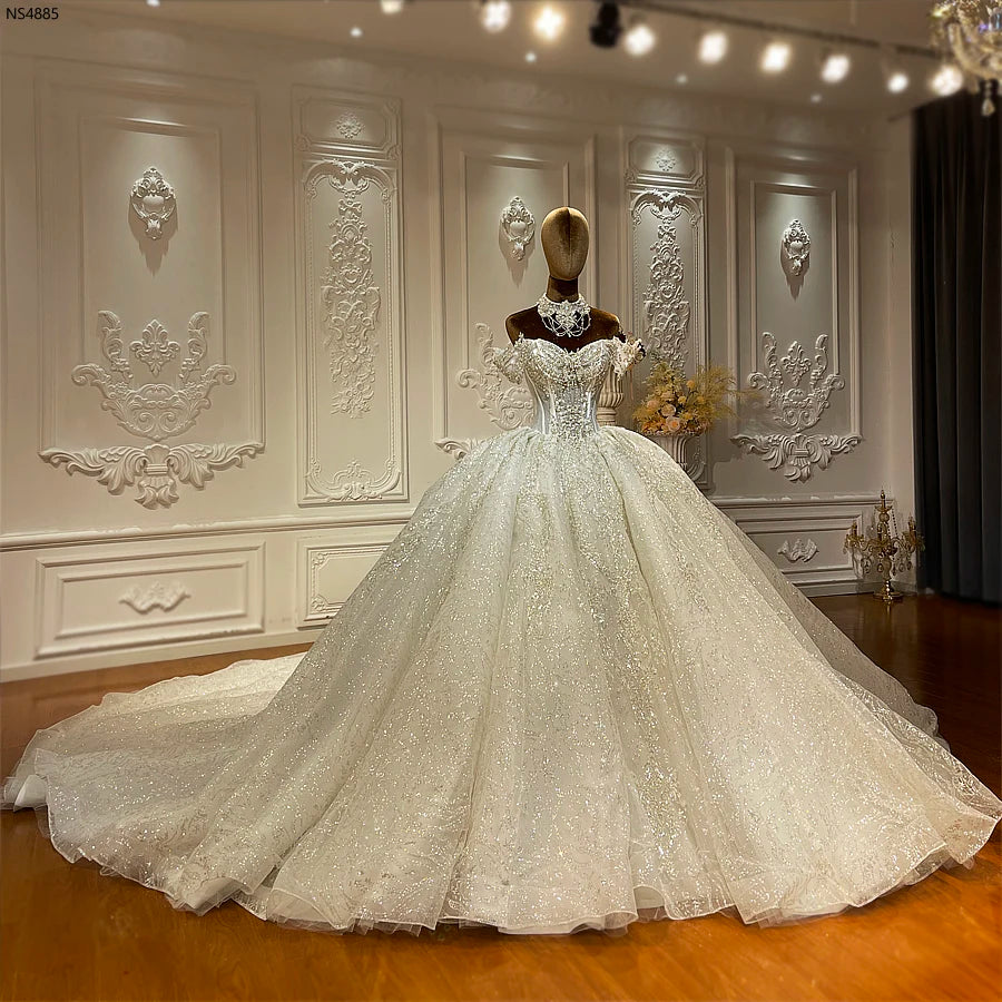 A luxurious, sparkling white ball gown on a mannequin stands in an ornate room with intricate wall moldings, exuding elegance and grandeur on a polished wooden floor.