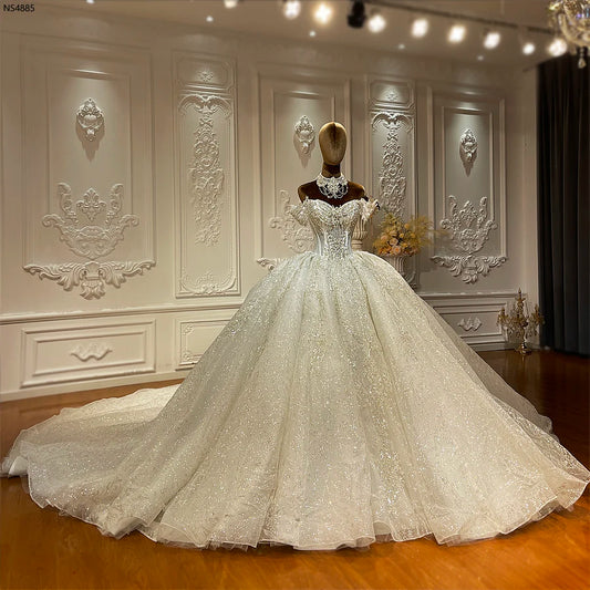 A luxurious, sparkling white ball gown on a mannequin stands in an ornate room with intricate wall moldings, exuding elegance and grandeur on a polished wooden floor.