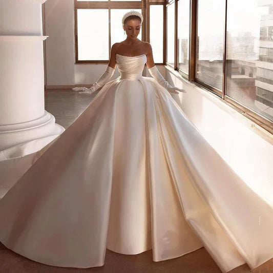 Bride in elegant off-the-shoulder satin wedding gown with fitted bodice and flowing skirt, wearing long gloves and a beaded headpiece, stands in sunlit room by large windows.