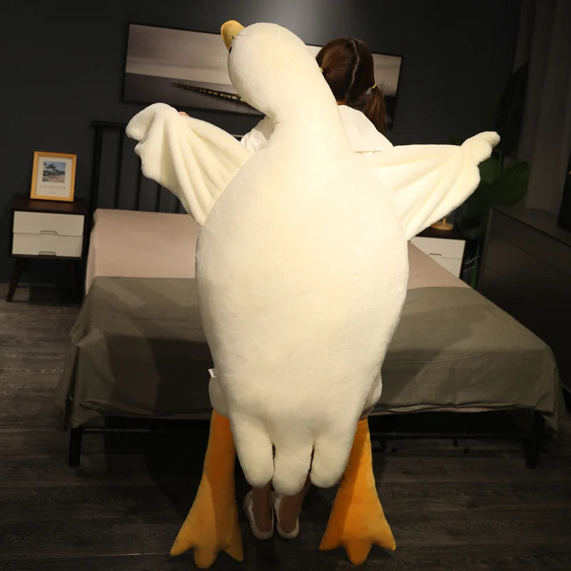 A person carries a large, fluffy white duck plushie with orange feet in a dimly lit bedroom. A bed with brown sheets and nightstand with a framed picture are in the background.