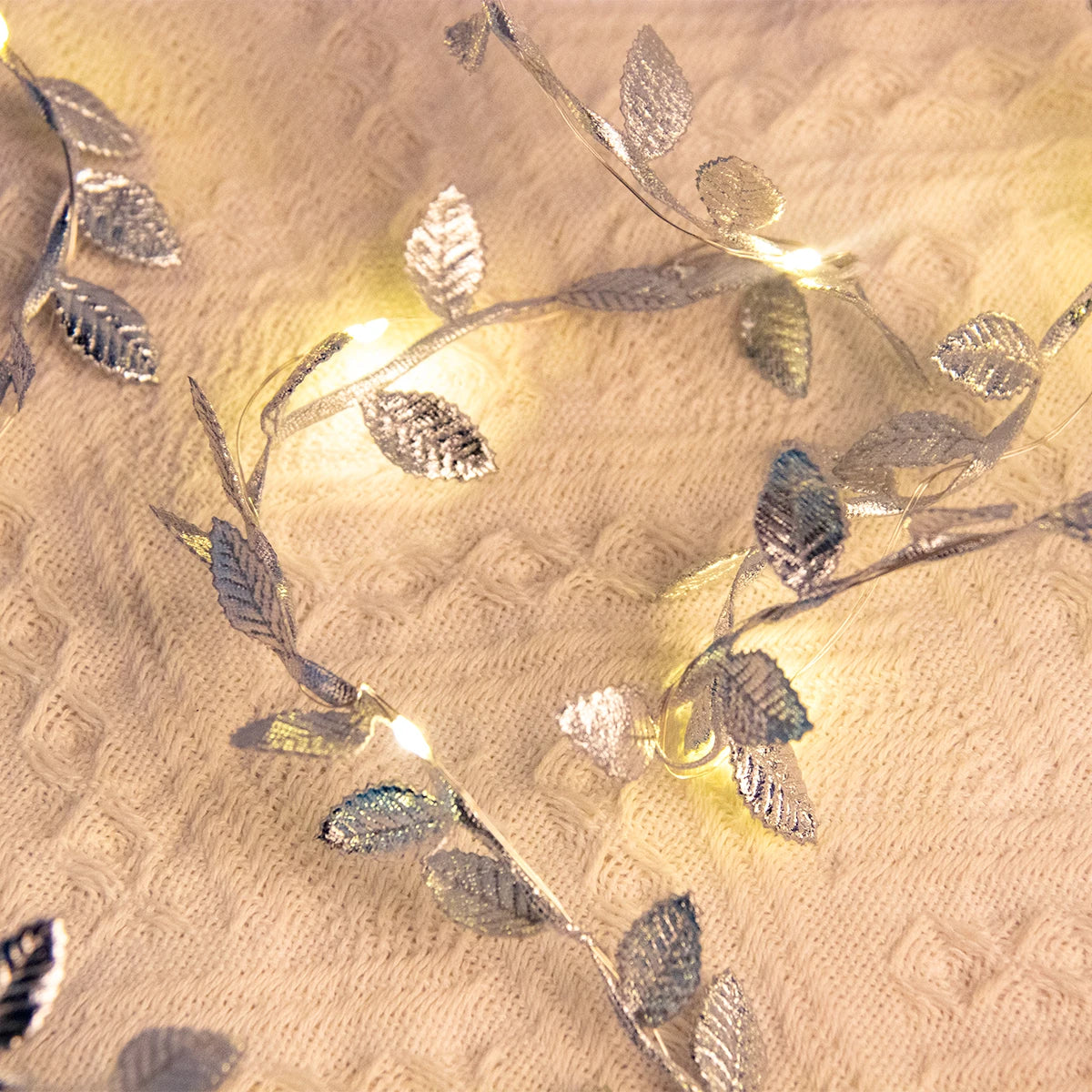Silver leaf string lights glow softly on a cream knit blanket, creating a cozy and festive ambiance. The metallic leaves shimmer in the warm light.