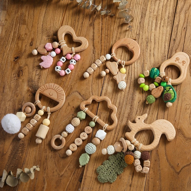 Wooden baby teething toys on a wooden surface, shaped like a heart, moon, sun, cloud, and squirrel. Each has colorful beads and crochet embellishments.