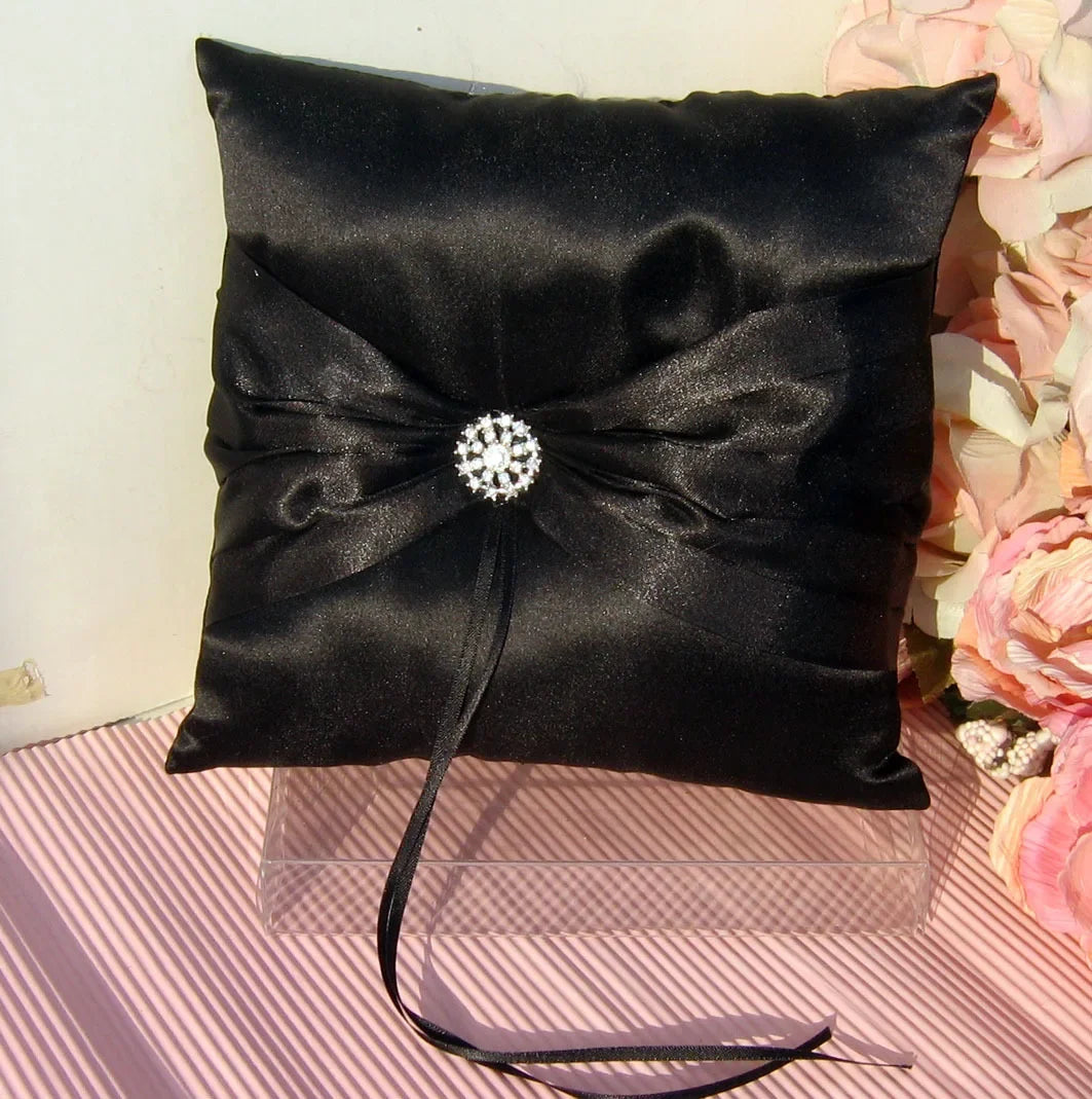 A black satin ring bearer pillow with a decorative crystal brooch at the center, placed on a striped surface with pink flowers in the background.