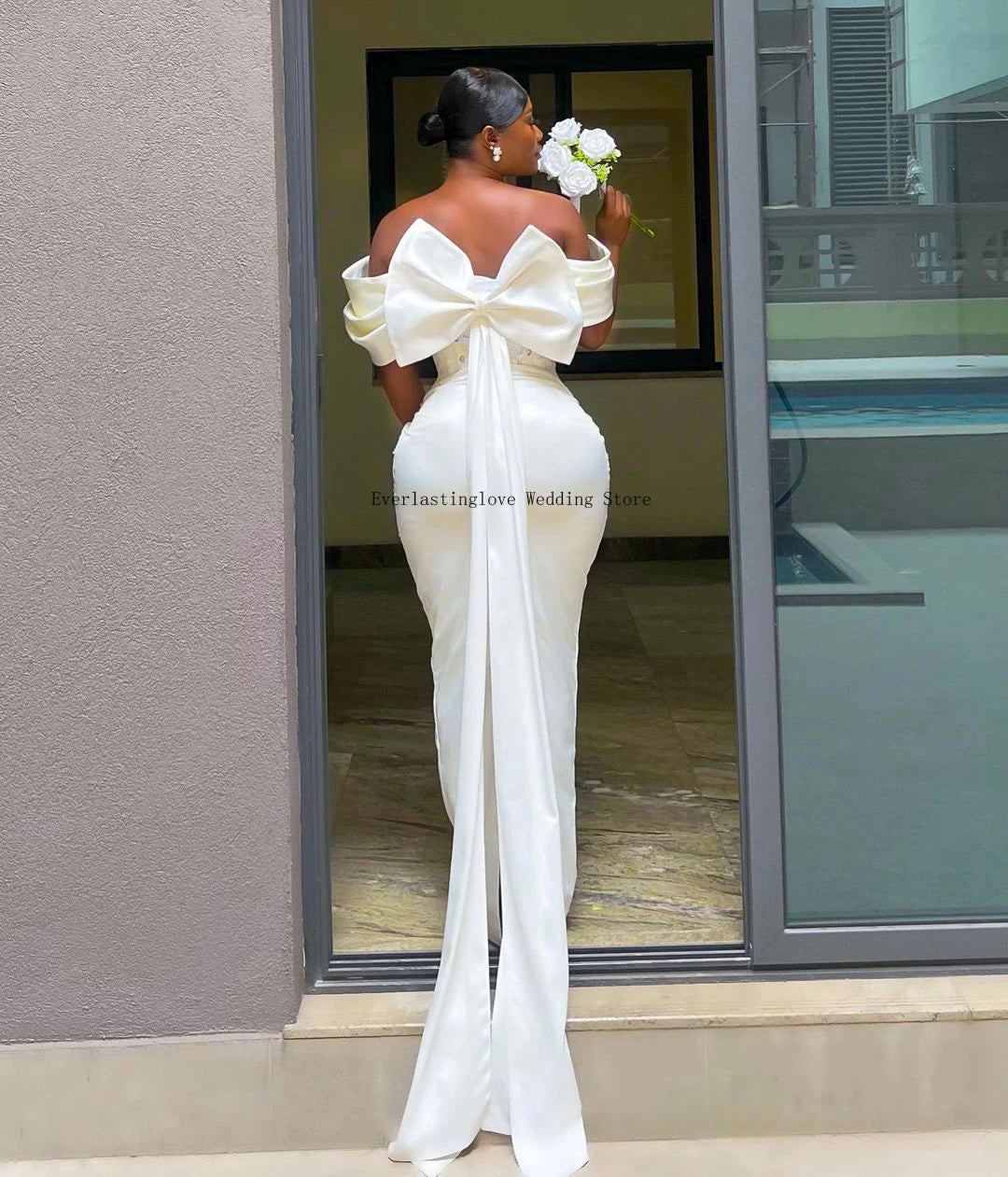 Woman in an elegant white gown holding a bouquet, with her back to the camera. The dress features an oversized bow and a long train. She stands by a glass door.