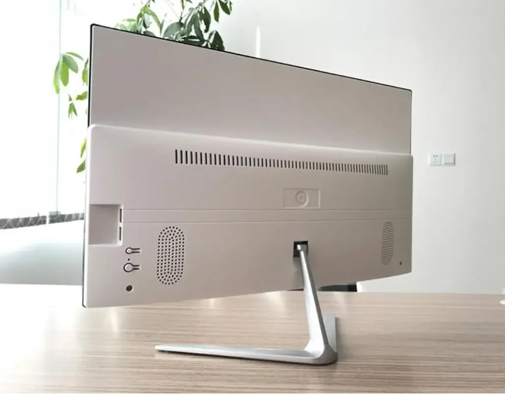 Back view of a sleek, slim monitor on a wooden desk, featuring ventilation slits and ports. Indoor setting with natural light and green plants in the background.