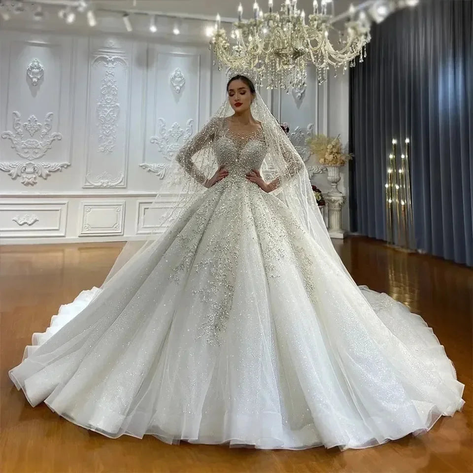 Elegant bride in a luxurious, sparkling wedding gown with intricate lace details, posing in an ornate room with chandeliers and elegant white paneling.