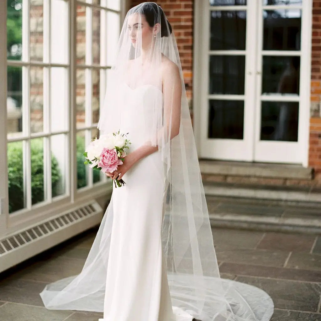 Bride in a white gown and long veil holding a bouquet of pink and white flowers. She stands indoors by large windows with a view of greenery and brick walls.