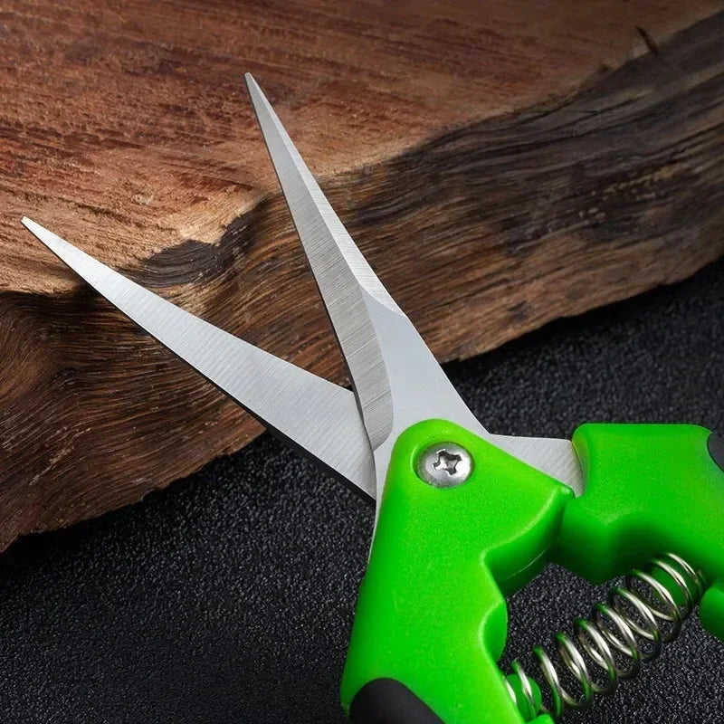Green-handled scissors with sharp metal blades, resting against a piece of textured wood on a black surface. A visible spring mechanism aids in opening the blades.