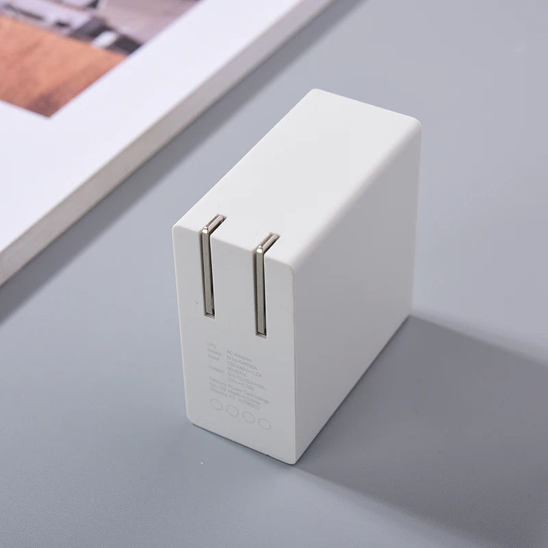 A white rectangular dual USB wall charger with two foldable prongs is placed on a gray surface. A corner of a framed picture is visible in the top left background.
