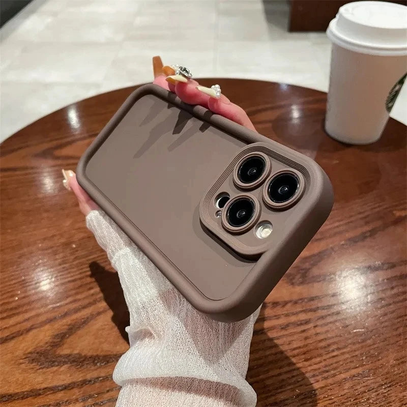 A hand with a white sleeve holds a smartphone with a brown case, showcasing three camera lenses, over a wooden table. A coffee cup is in the background.