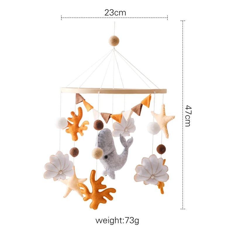 A 47cm baby mobile featuring a gray whale, orange seaweed, starfish, and felt balls, hanging from a wooden ring. It weighs 73g and is 23cm wide.
