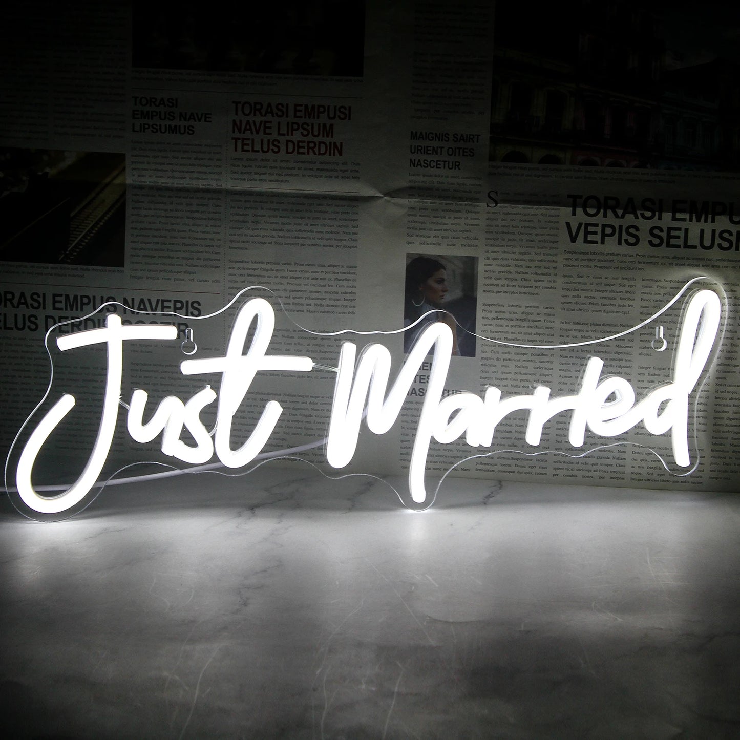 Illuminated "Just Married" sign with bright white neon letters in script font, set against a dark, newspaper-covered background.