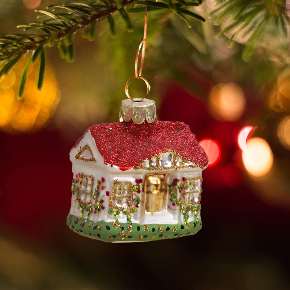 Festive ornament shaped like a house with a sparkling red roof, hanging from a Christmas tree branch. The background is warmly lit with a holiday ambiance.