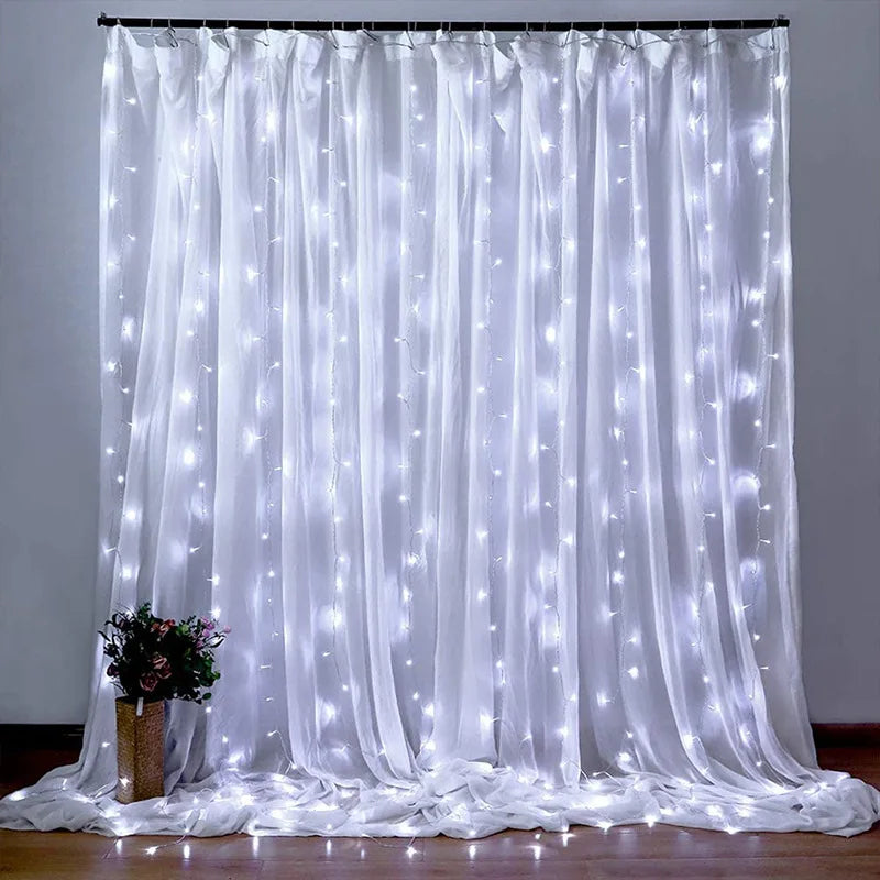White sheer curtains adorned with cascading string lights create a softly lit backdrop. A vase filled with flowers sits on the floor to the left, adding a decorative touch.