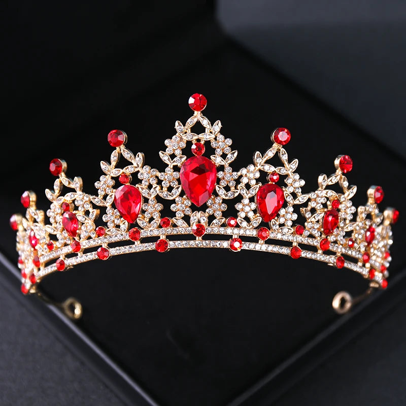 A gold tiara adorned with sparkling red and clear gemstones arranged in intricate floral patterns, set against a dark background.