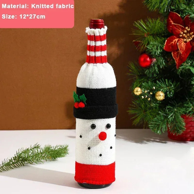 A wine bottle wrapped in a snowman-themed knitted cover with a red, black, and white design, small buttons, and a green holly decoration. A Christmas tree is in the background.