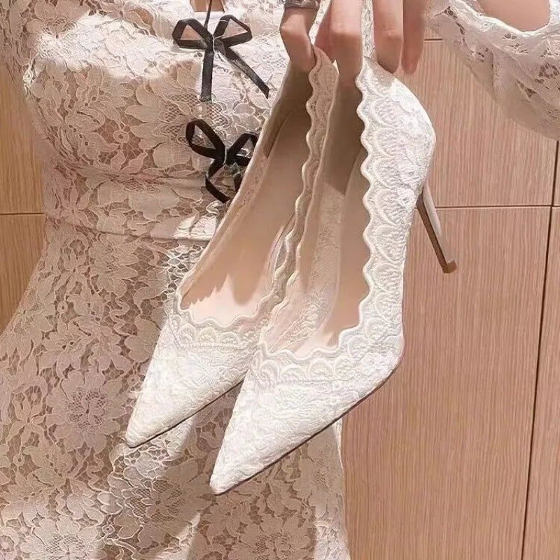 A person holds a pair of elegant white lace stiletto heels with scalloped edges, matching their lace dress adorned with black bows.