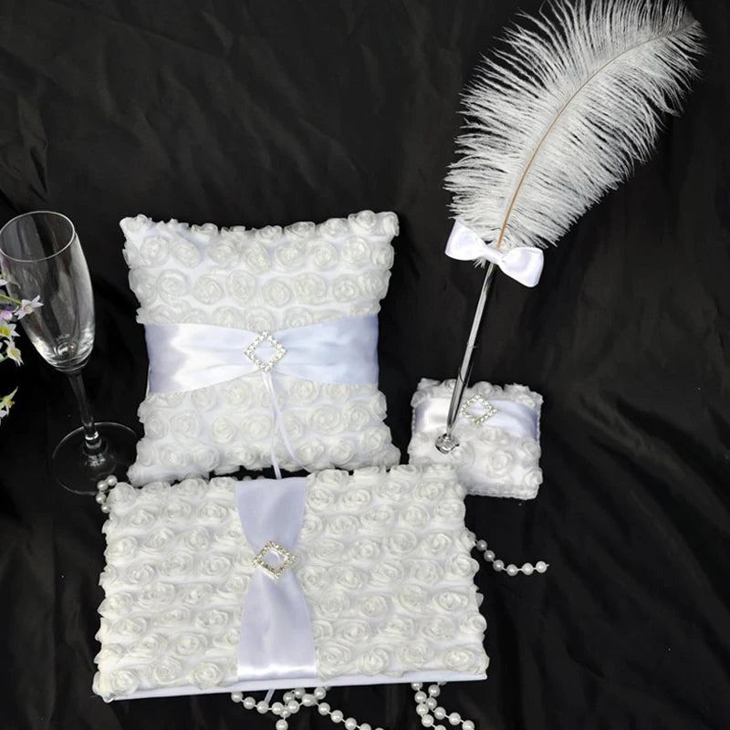Elegant wedding set with white floral-patterned pillow, pen with feather and bow, guest book, ring pillow, champagne flute, and pearl beads on a black background.