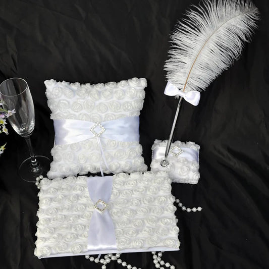 Elegant wedding set with white floral-patterned pillow, pen with feather and bow, guest book, ring pillow, champagne flute, and pearl beads on a black background.