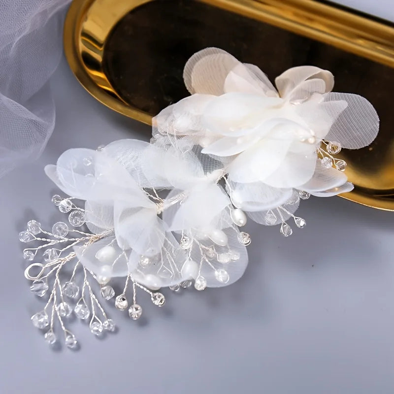 Delicate white floral hair accessory with chiffon petals, pearls, and crystal beads, placed on a reflective gold tray on a soft gray background.