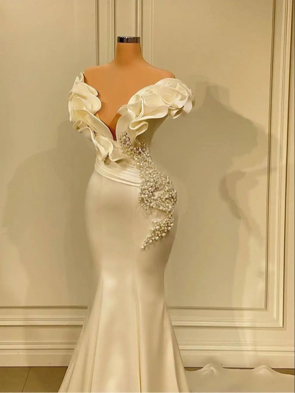 Elegant off-shoulder white gown on mannequin, featuring floral ruffles and pearl embellishments on the bodice, flaring into a mermaid-style skirt.