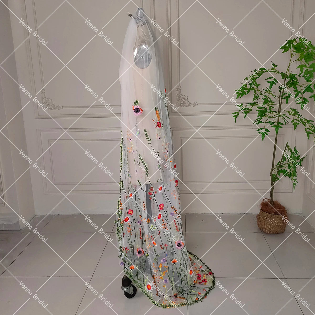A mannequin displays a sheer bridal veil adorned with colorful floral embroidery. The setup is in a minimalistic room with a potted plant in the corner.