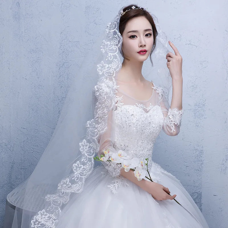 Bride in an elegant white lace wedding dress holding a white flower. The veil features intricate floral lace, and her hair is adorned with delicate accessories.