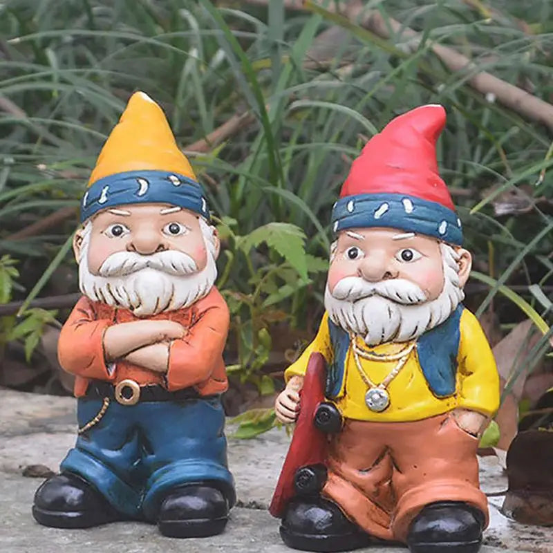 Two colorful garden gnomes: one in an orange hat and crossed arms, the other in a red hat holding a skateboard. They stand on stone with greenery in the background.